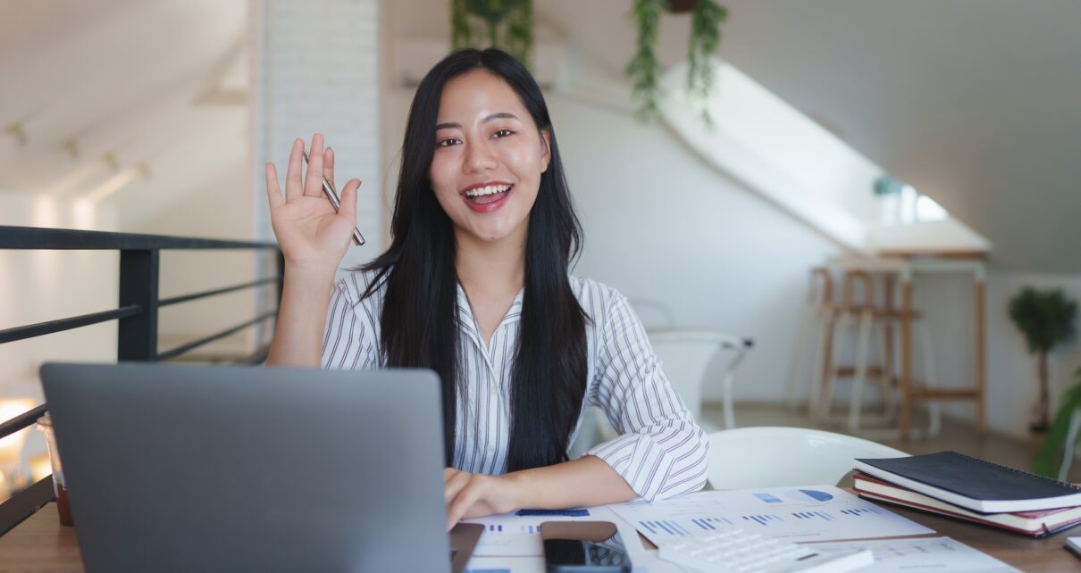 Asian businesswoman waving at someone during a video call in her bright office
