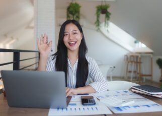 Asian businesswoman waving at someone during a video call in her bright office