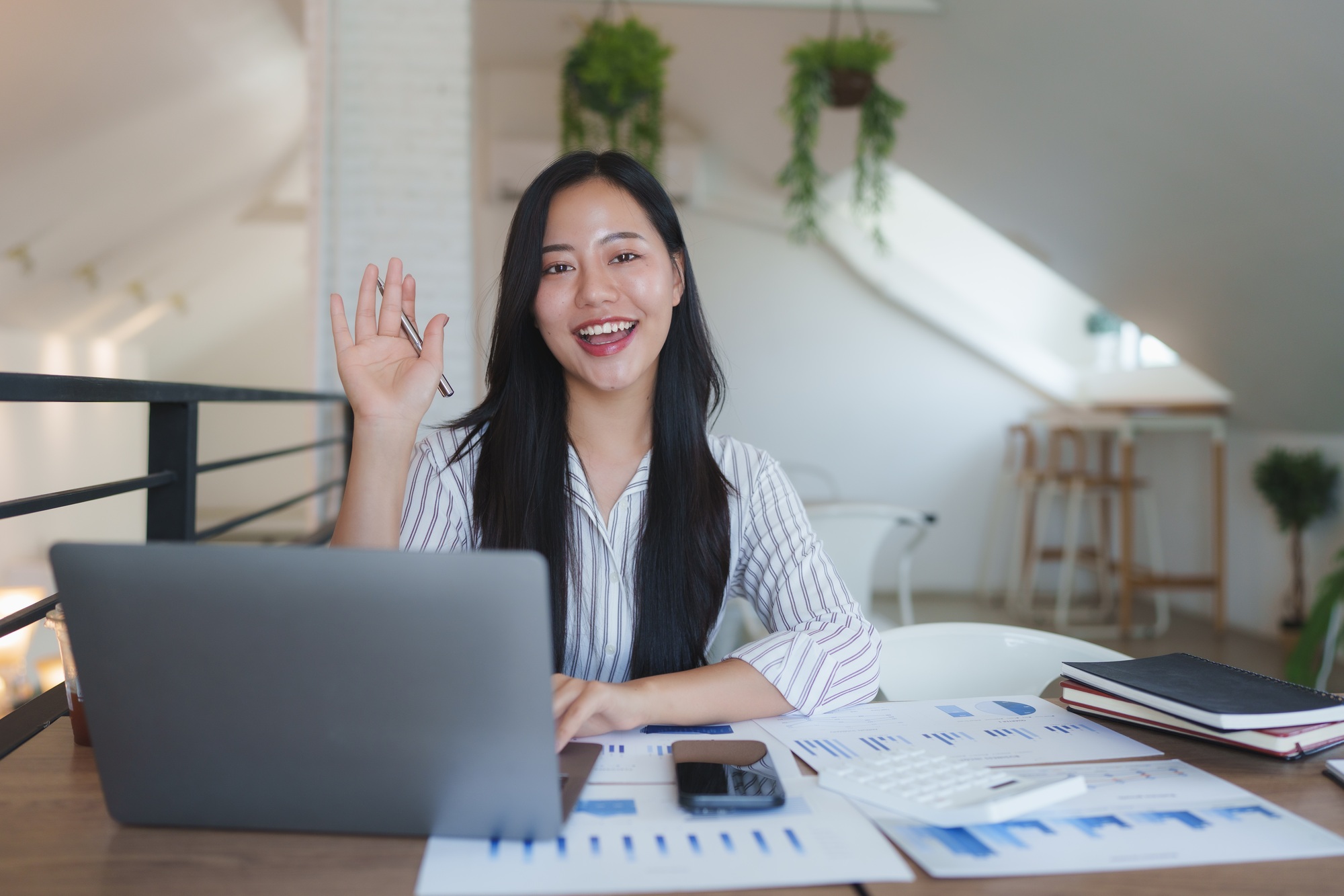 Asian businesswoman waving at someone during a video call in her bright office
