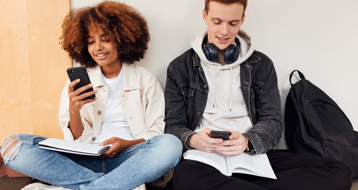 Classmates sitting at wall in school holding smartphones
