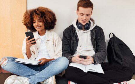 Classmates sitting at wall in school holding smartphones