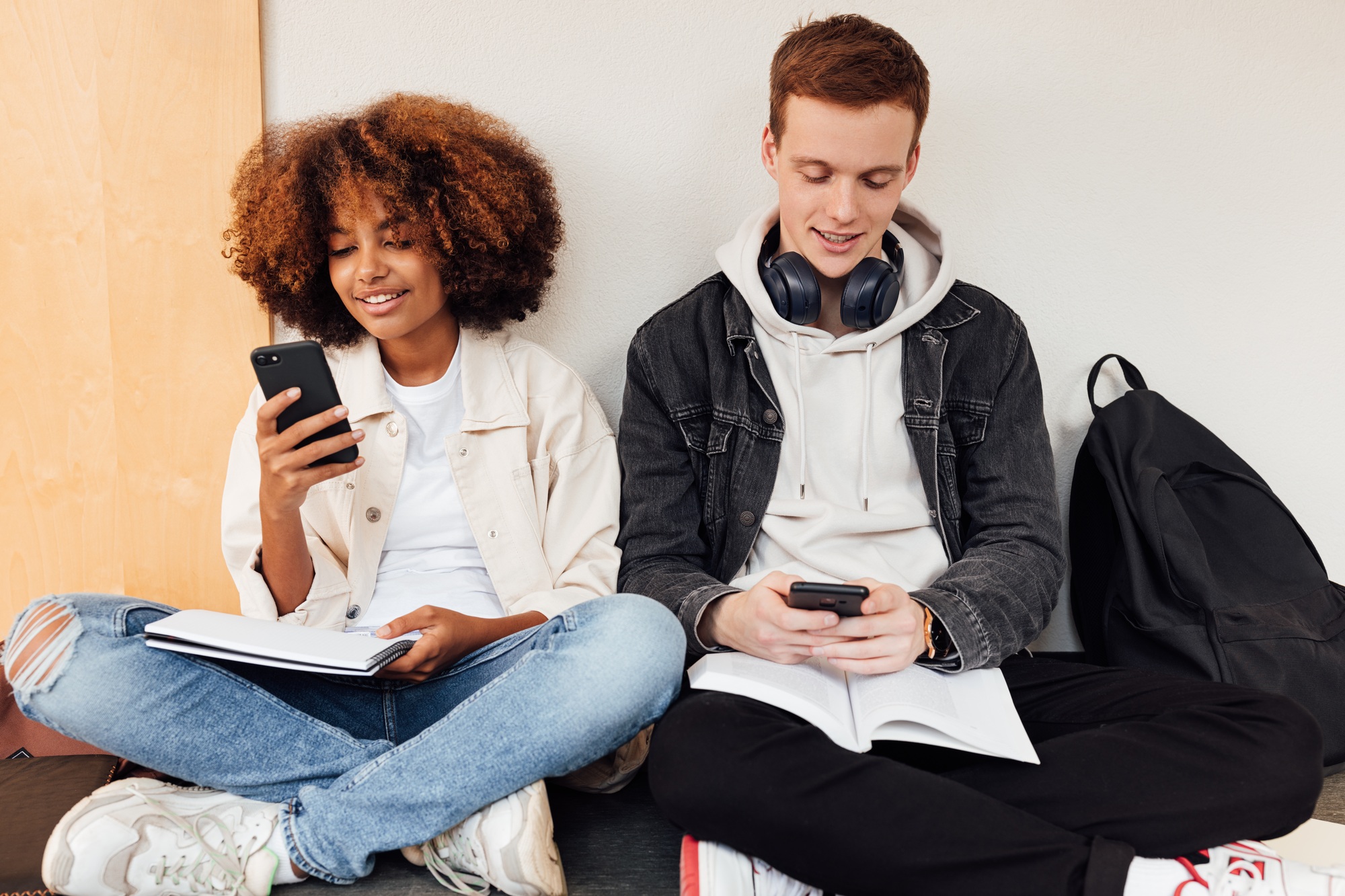Classmates sitting at wall in school holding smartphones